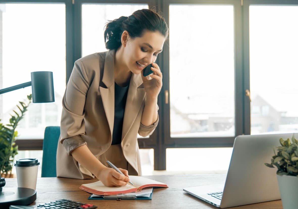 A woman speaks on the phone while writing in an open planner beside a laptop.